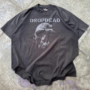 May include: A charcoal gray short-sleeve t-shirt featuring the text DROPDEAD in a distressed white font above a graphic illustration of a skull. The shirt has a crew neckline and a standard fit, showcasing a faded, worn-in appearance throughout.