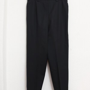 May include: Black trousers hanging on a wooden clothes hanger. The trousers have a straight leg design and are made of a solid black fabric. The trousers are hanging against a white wall.