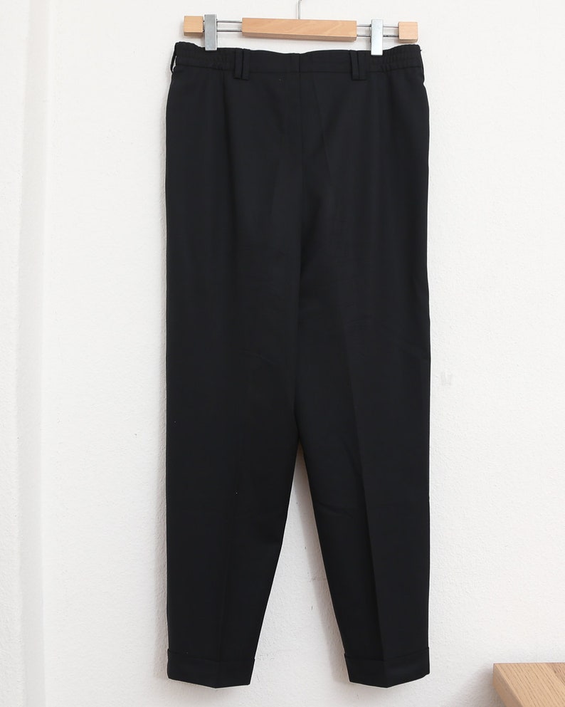May include: Black trousers hanging on a wooden clothes hanger. The trousers have a straight leg design and are made of a solid black fabric. The trousers are hanging against a white wall.