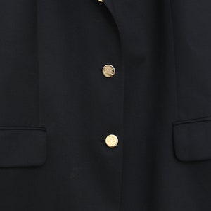 May include: A close-up of a black blazer with two gold buttons and two front pockets. The buttons have a circular design. The blazer is made of a smooth fabric.