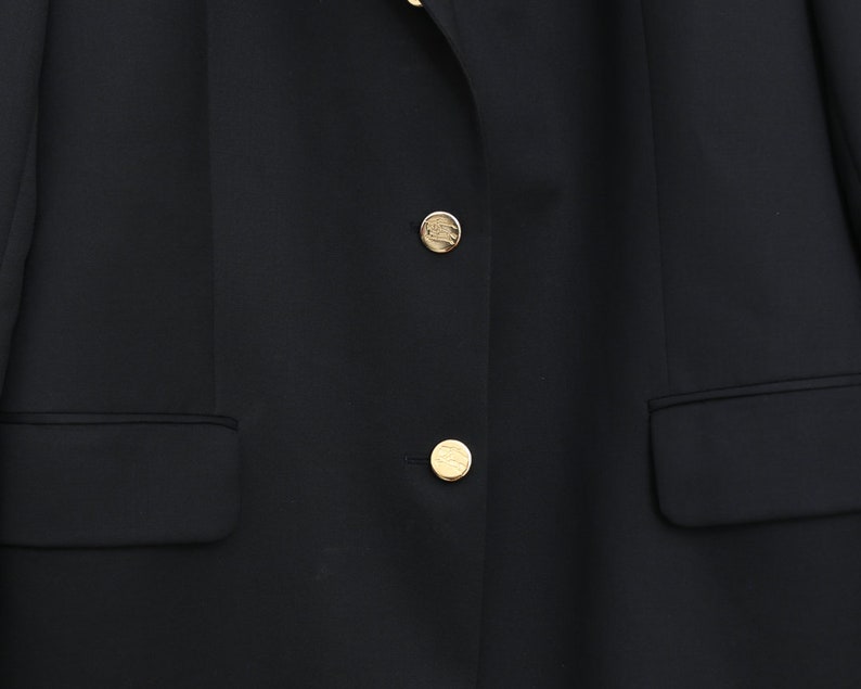 May include: A close-up of a black blazer with two gold buttons and two front pockets. The buttons have a circular design. The blazer is made of a smooth fabric.