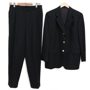 May include: A black suit comprising a blazer and trousers, displayed against a white backdrop. The blazer has gold-coloured buttons and a tailored design. The trousers are pleated with a straight leg. The suit is on a wooden hanger.