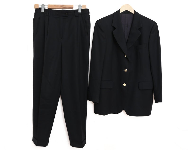 May include: A black suit comprising a blazer and trousers, displayed against a white backdrop. The blazer has gold-coloured buttons and a tailored design. The trousers are pleated with a straight leg. The suit is on a wooden hanger.