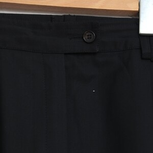May include: Close-up of black trousers with a button closure and belt loops. The fabric is a smooth, solid black. The trousers are hanging on a wooden hanger against a white brick wall.
