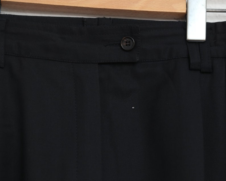 May include: Close-up of black trousers with a button closure and belt loops. The fabric is a smooth, solid black. The trousers are hanging on a wooden hanger against a white brick wall.