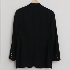 May include: A black blazer with gold buttons on the sleeves, hanging on a wooden hanger. The blazer is against a white wall. The design is classic and tailored. The buttons are gold-coloured.