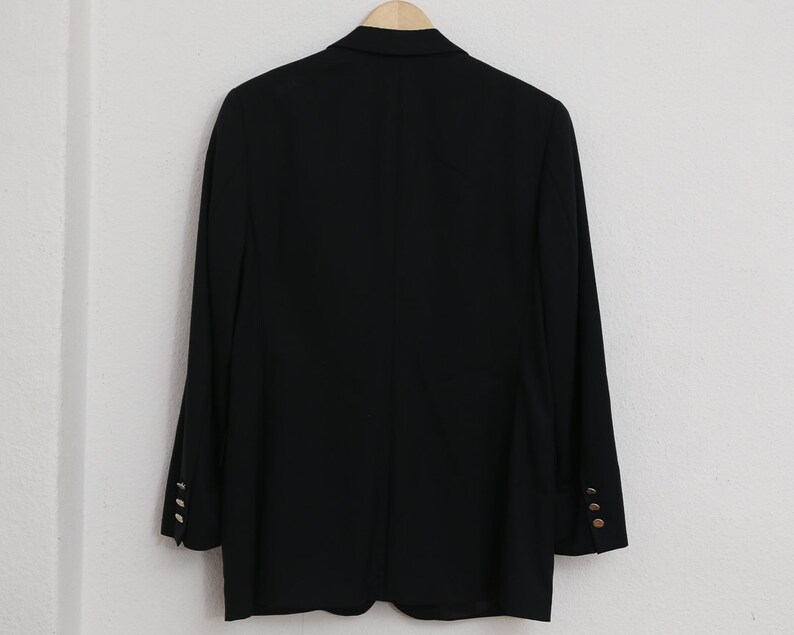 May include: A black blazer with gold buttons on the sleeves, hanging on a wooden hanger. The blazer is against a white wall. The design is classic and tailored. The buttons are gold-coloured.