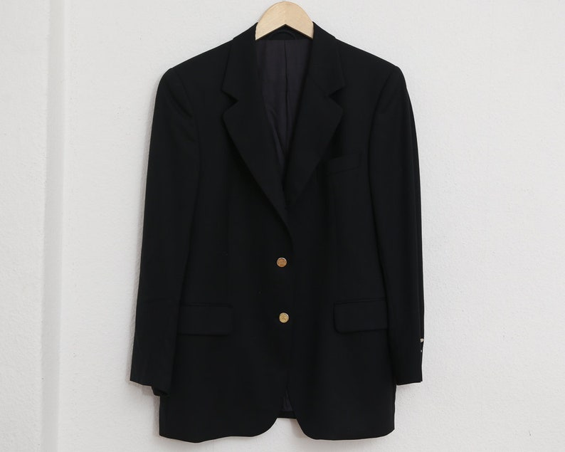 May include: A black blazer with gold buttons hangs on a wooden hanger. The blazer features a classic design with a notched lapel and two front pockets. The gold buttons are visible on the front of the blazer.