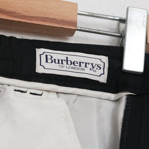 May include: Close-up of black trousers with a white lining. The label reads "Burberrys of London" in a navy blue frame. The trousers are hanging on a wooden hanger with a metal clip.