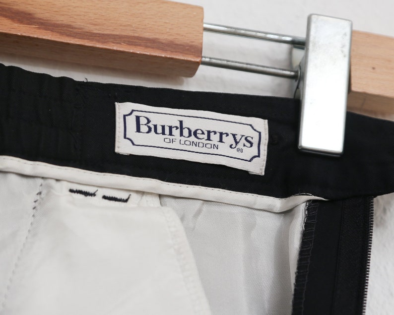 May include: Close-up of black trousers with a white lining. The label reads "Burberrys of London" in a navy blue frame. The trousers are hanging on a wooden hanger with a metal clip.