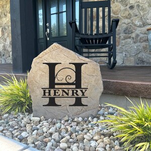 May include: A personalized stone with the letter "H" and the name "HENRY" engraved on it. The stone is sitting on a bed of small rocks in front of a house.
