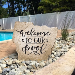 May include: A large, light brown stone with the words "welcome to our pool" written in black lettering. The stone is sitting on a bed of gray and white pebbles in front of a white fence and a swimming pool.