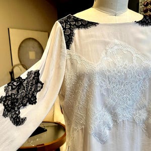 May include: White blouse with long sleeves, featuring intricate black lace detailing on the shoulders and sleeves. The bodice showcases delicate white lace patterns. The garment is displayed on a mannequin, highlighting its design and texture.