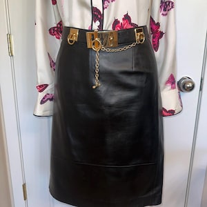May include: A black leather skirt with a gold-tone belt and chain detail. The skirt is styled with a white blouse patterned with pink butterflies. The outfit is displayed on a mannequin.
