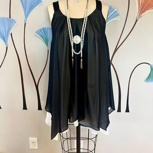 May include: A black sleeveless top with a white underlay, displayed on a mannequin. A long pearl necklace with a decorative pendant and tassel accents is draped over the top. The background features a wall with colorful floral art.