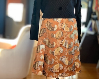 May include: A black turtleneck sweater with a cable knit pattern and black pom pom buttons. The sweater is paired with a brown skirt with a gold and silver paisley print.