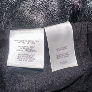 May include: Close-up of a black leather item with a textured surface. Two white tags are visible, one with "PROFESSIONAL DRY CLEAN LEATHER METHOD" and the other with "100% LEATHER" and "MADE IN CHINA".