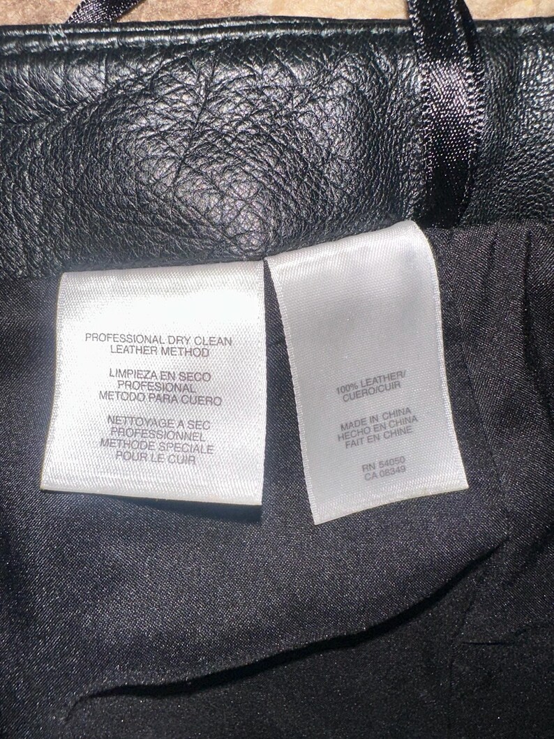 May include: Close-up of a black leather item with a textured surface. Two white tags are visible, one with "PROFESSIONAL DRY CLEAN LEATHER METHOD" and the other with "100% LEATHER" and "MADE IN CHINA".