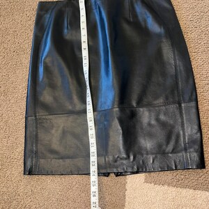 May include: A black leather skirt with a straight cut, measured with a white measuring tape. The skirt appears to be approximately 71 cm long. The skirt has a classic design and is made of a smooth, dark material.