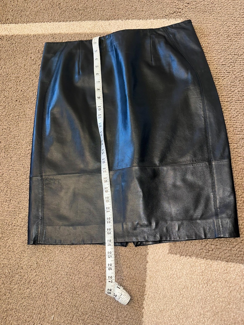 May include: A black leather skirt with a straight cut, measured with a white measuring tape. The skirt appears to be approximately 71 cm long. The skirt has a classic design and is made of a smooth, dark material.