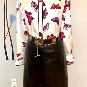 May include: A white blouse with a black collar and trim, patterned with pink and purple butterflies. A black leather skirt is worn with a gold-tone belt featuring a chain detail.