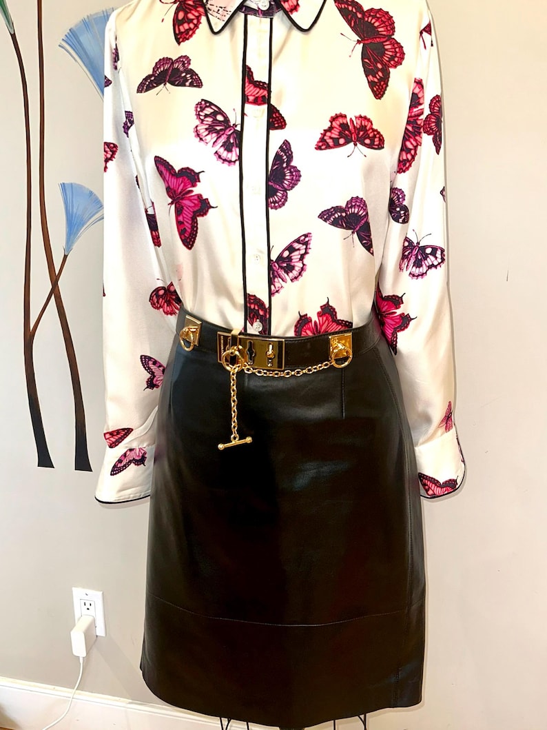 May include: A white blouse with a black collar and trim, patterned with pink and purple butterflies. A black leather skirt is worn with a gold-tone belt featuring a chain detail.