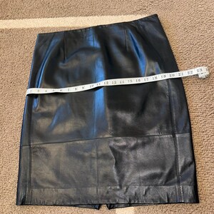 May include: A black leather skirt with a straight cut, featuring a back slit and visible stitching details. The skirt is measured with a tape measure, showing a length of approximately 53 cm.