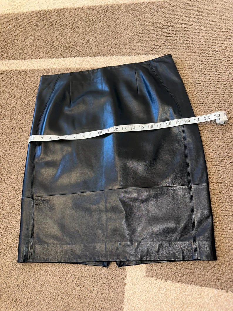 May include: A black leather skirt with a straight cut, featuring a back slit and visible stitching details. The skirt is measured with a tape measure, showing a length of approximately 53 cm.
