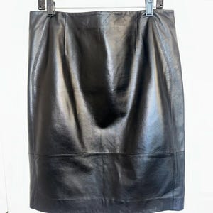 May include: A black leather skirt hanging on a black hanger. The skirt is knee-length with a straight silhouette. The leather has a glossy finish and visible stitching along the seams. The skirt appears to be a classic, versatile piece.
