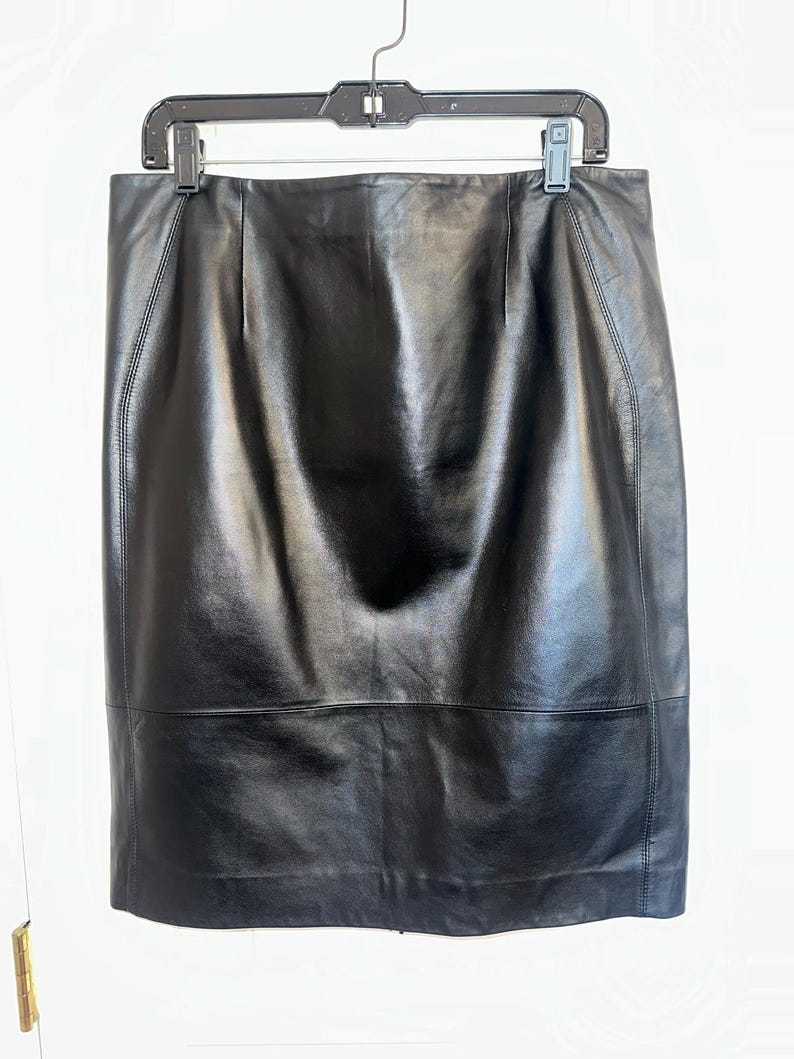 May include: A black leather skirt hanging on a black hanger. The skirt is knee-length with a straight silhouette. The leather has a glossy finish and visible stitching along the seams. The skirt appears to be a classic, versatile piece.