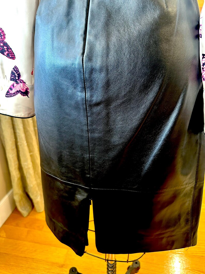 May include: A black leather skirt with a split at the back hem, displayed on a mannequin. A white blouse with butterfly patterns is partially visible on the left. The skirt is made of leather.