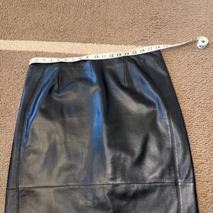 May include: Black leather skirt with a straight cut. A measuring tape is laid across the waist, showing a measurement. The skirt appears to be made of smooth leather. The skirt is on a brown carpet.
