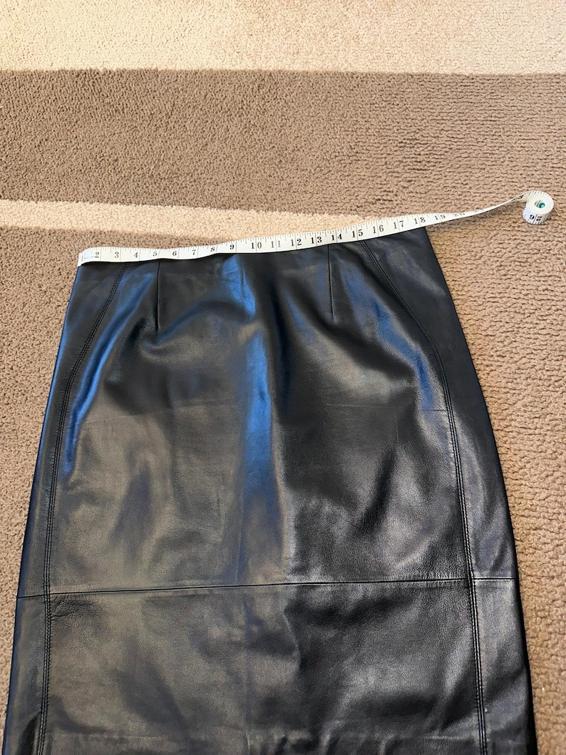 May include: Black leather skirt with a straight cut. A measuring tape is laid across the waist, showing a measurement. The skirt appears to be made of smooth leather. The skirt is on a brown carpet.