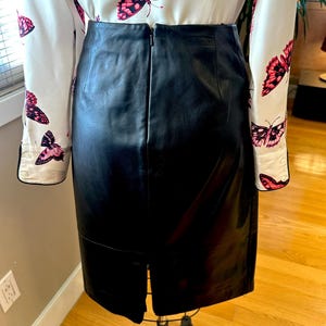 May include: A black leather skirt with a back zip and a hem slit, displayed on a mannequin. A white blouse with a pink butterfly pattern is partially visible.