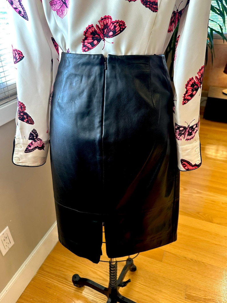 May include: A black leather skirt with a back zip and a hem slit, displayed on a mannequin. A white blouse with a pink butterfly pattern is partially visible.