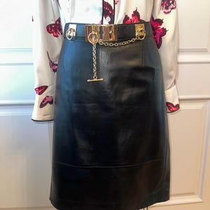 May include: A black leather skirt with a gold chain belt. The belt has a rectangular buckle and a dangling chain with a T-bar closure. The skirt is paired with a white blouse featuring a butterfly pattern in shades of pink and red.