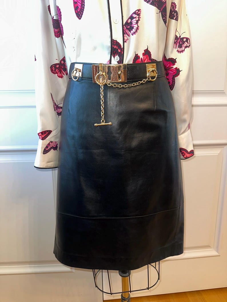 May include: A black leather skirt with a gold chain belt. The belt has a rectangular buckle and a dangling chain with a T-bar closure. The skirt is paired with a white blouse featuring a butterfly pattern in shades of pink and red.