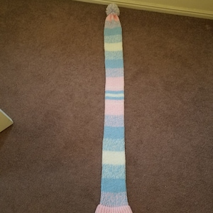 May include: A long, striped scarf with a pom-pom on top. The scarf is made of a soft, knitted fabric and features alternating stripes of pink, light blue, and white.
