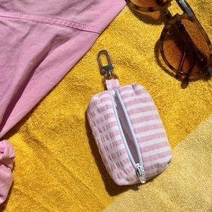 Può includere: Una piccola custodia rettangolare in tessuto a righe rosa e bianche, dotata di un moschettone in metallo argentato e di una chiusura a zip bianca. La custodia è parzialmente aperta, rivelando una fodera interna bianca e una consistenza morbida.