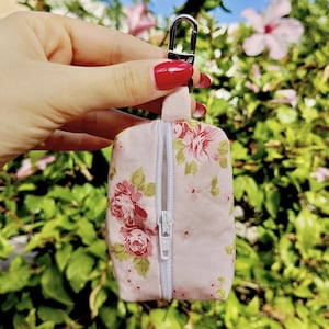 Puede incluir: Una pequeña bolsa rectangular de tela con fondo rosa claro y un estampado floral de rosas rojas y hojas verdes. La bolsa tiene una cremallera vertical blanca en el centro y un mosquetón de metal plateado sujeto en la parte superior.