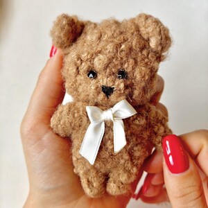Op de afbeelding: Een kleine, lichtbruine teddybeer gemaakt van getextureerd, krullend garen. De beer heeft twee ronde oren, kleine zwarte kraaloogjes, een geborduurde zwarte neus en een wit satijnen lint dat als een strik om de nek is gebonden. Het oppervlak is pluizig.