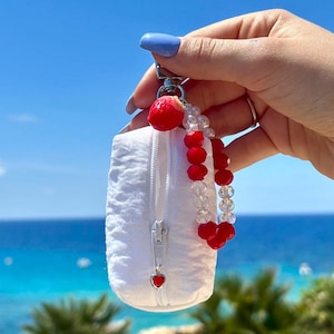 Könnte beinhalten: Eine weiße, gesteppte Stofftasche mit silbernem Reißverschluss und herzförmigem Anhänger. Sie ist an einem silbernen Verschluss mit rotem Perlenakzent und einer Kette aus roten und klaren Perlen befestigt. Der Hintergrund ist ein blauer Himmel und das Meer.