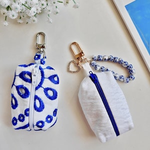 May include: Two small rectangular zippered pouches. The left pouch is white with blue embroidered eyelet patterns and a silver swivel clasp. The right pouch is plain white with a blue zipper, a blue and white floral beaded wrist strap, and a heart charm.