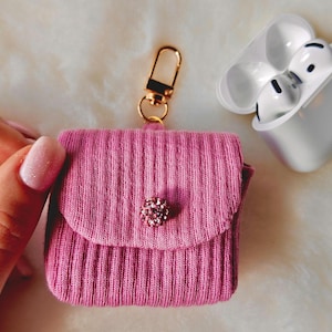 May include: A pink ribbed knit AirPod case with a gold-tone clasp and a decorative jewel. The case is next to a white AirPod charging case with white earbuds.
