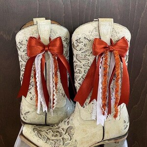 Op de afbeelding: Een paar cr&egrave;mekleurige cowboylaarzen met ingewikkelde bloempatronen. Elke laars is versierd met een roestoranje strik, wit kant en oranje tinsel. De laarzen hebben een klassiek western design met een puntige neus en een blokhak.