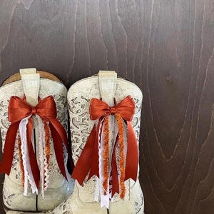 Op de afbeelding: Een paar off-white cowboylaarzen met ingewikkelde bloempatronen. Elke laars is versierd met een verbrande oranje strik en bijpassende linten, wit kant en oranje tinsel. De laarzen staan tegen een donkere houten achtergrond.