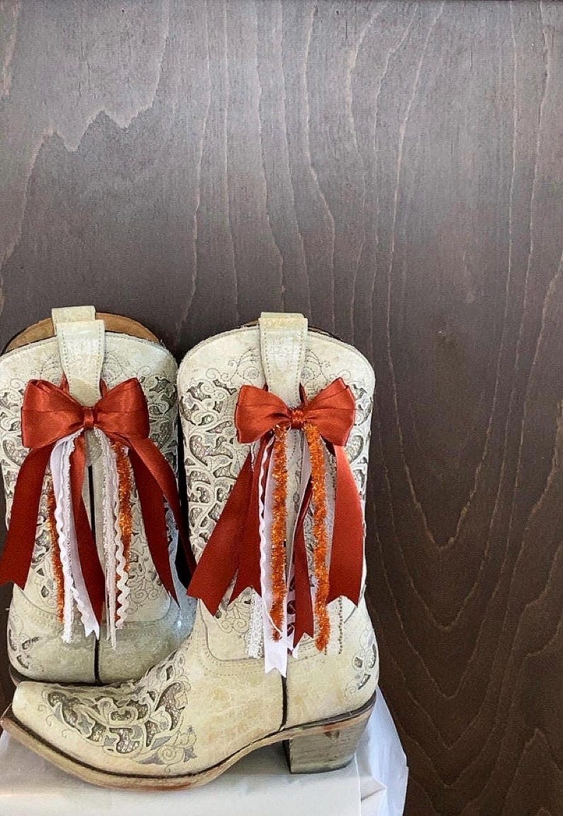 Op de afbeelding: Een paar off-white cowboylaarzen met ingewikkelde bloempatronen. Elke laars is versierd met een verbrande oranje strik en bijpassende linten, wit kant en oranje tinsel. De laarzen staan tegen een donkere houten achtergrond.