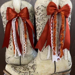 Op de afbeelding: Twee off-white cowboylaarzen met zilveren bloemmotieven. Elke laars heeft een roestkleurige strik, witte kant en oranje slingers. De laarzen hebben een klassiek western design met een puntige neus en een blokhak.