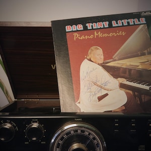 May include: Vintage record album titled "Big Tiny Little Piano Memories" with a photo of a man playing the piano, resting on top of a vintage radio. The album cover is signed. The radio is black with tuning dials.