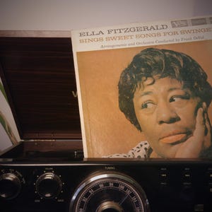 May include: Vintage vinyl record album cover featuring Ella Fitzgerald, titled "Sings Sweet Songs for Swingers." The album is set against a dark wooden record player, with the text visible on the album cover.